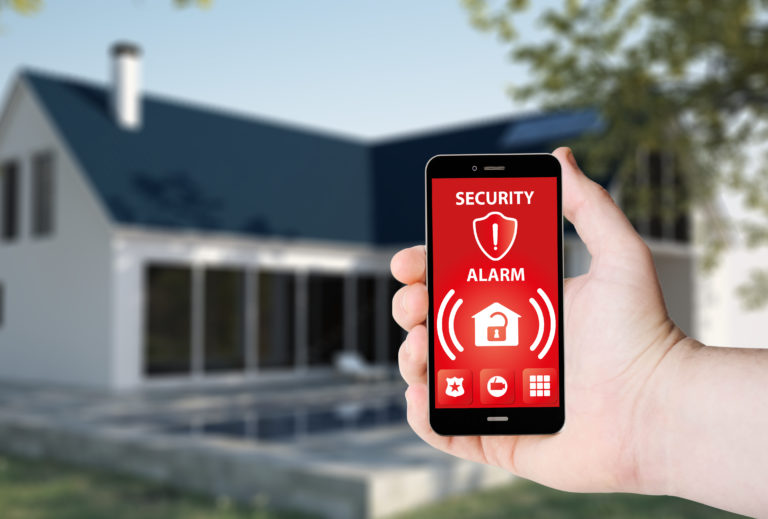 Hand hold a phone with security alarm app on a screen on the background of a house. All screen graphics are made up.